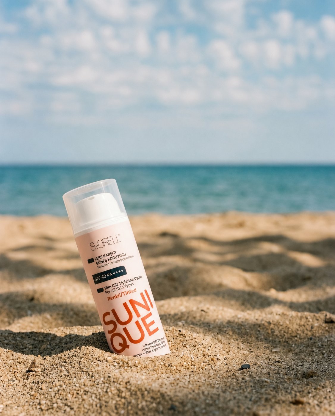 Sunique Spot Defence Tinted SPF 40 sunscreen bottle on warm dappled sand under scattered clouds