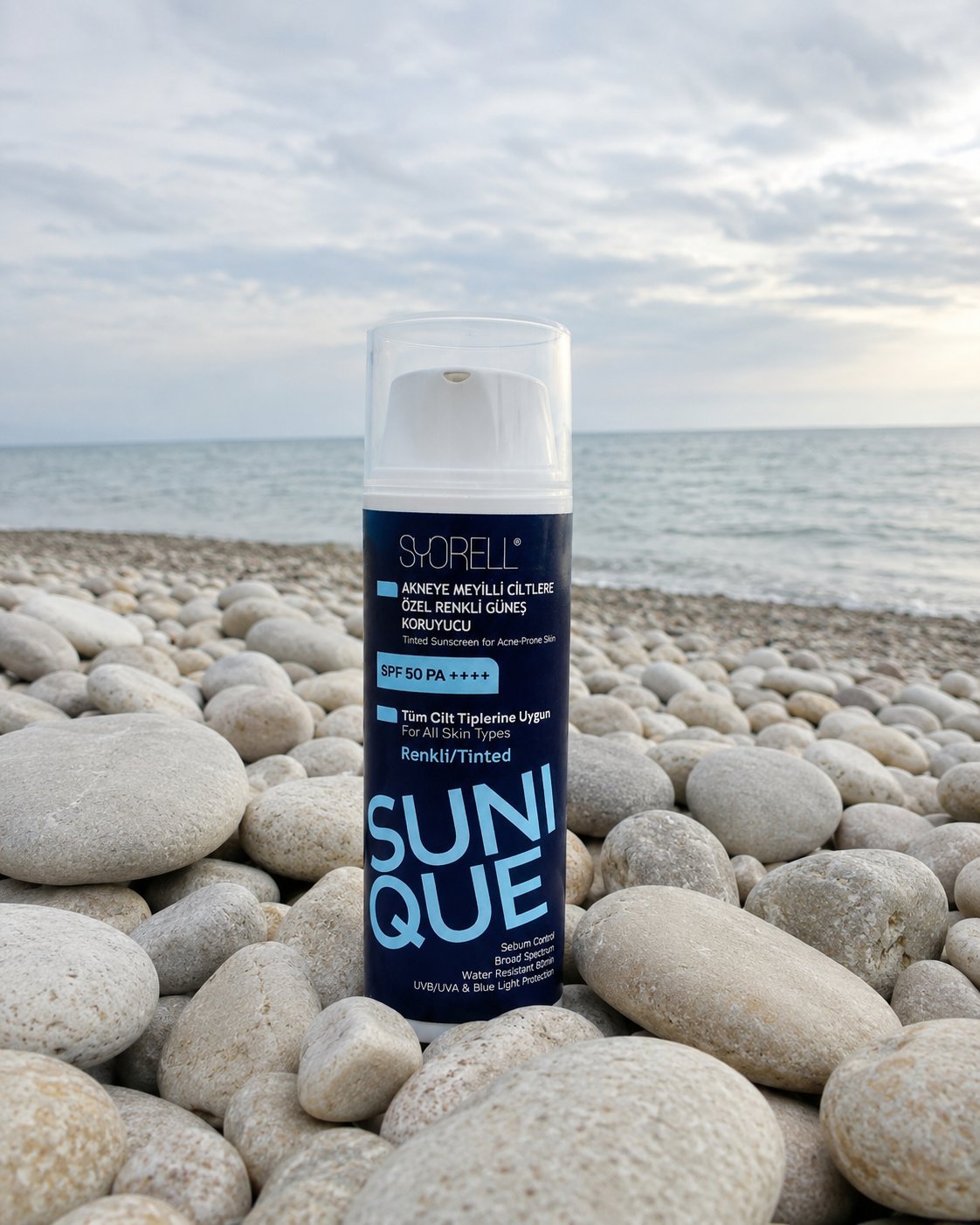 Sunique Acne-Prone Tinted SPF 50 sunscreen bottle on bone-white pebble shore at hazy summer morning