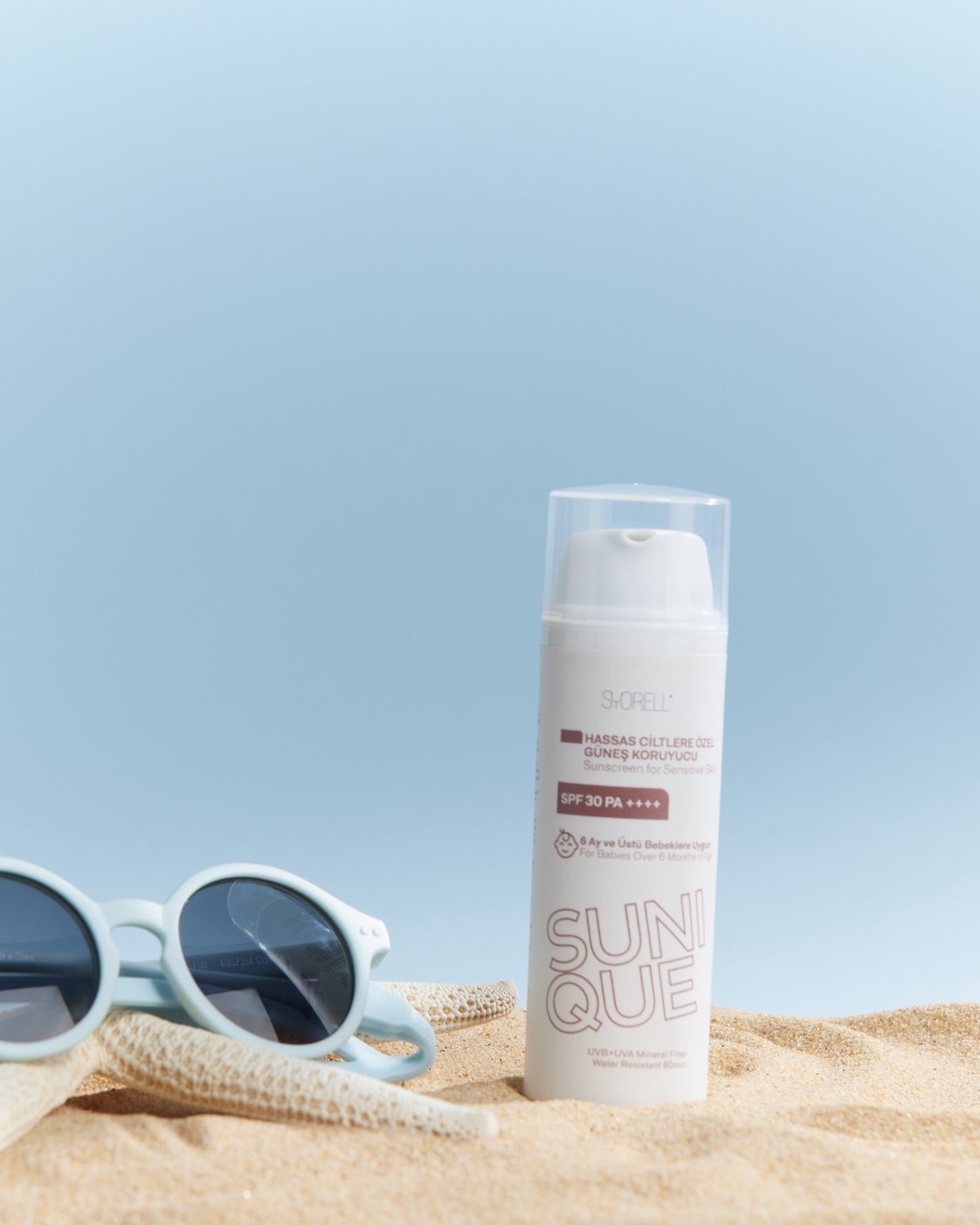 Sunique Sensitive Skin SPF 30 sunscreen bottle on soft sand with pale blue sunglasses and starfish