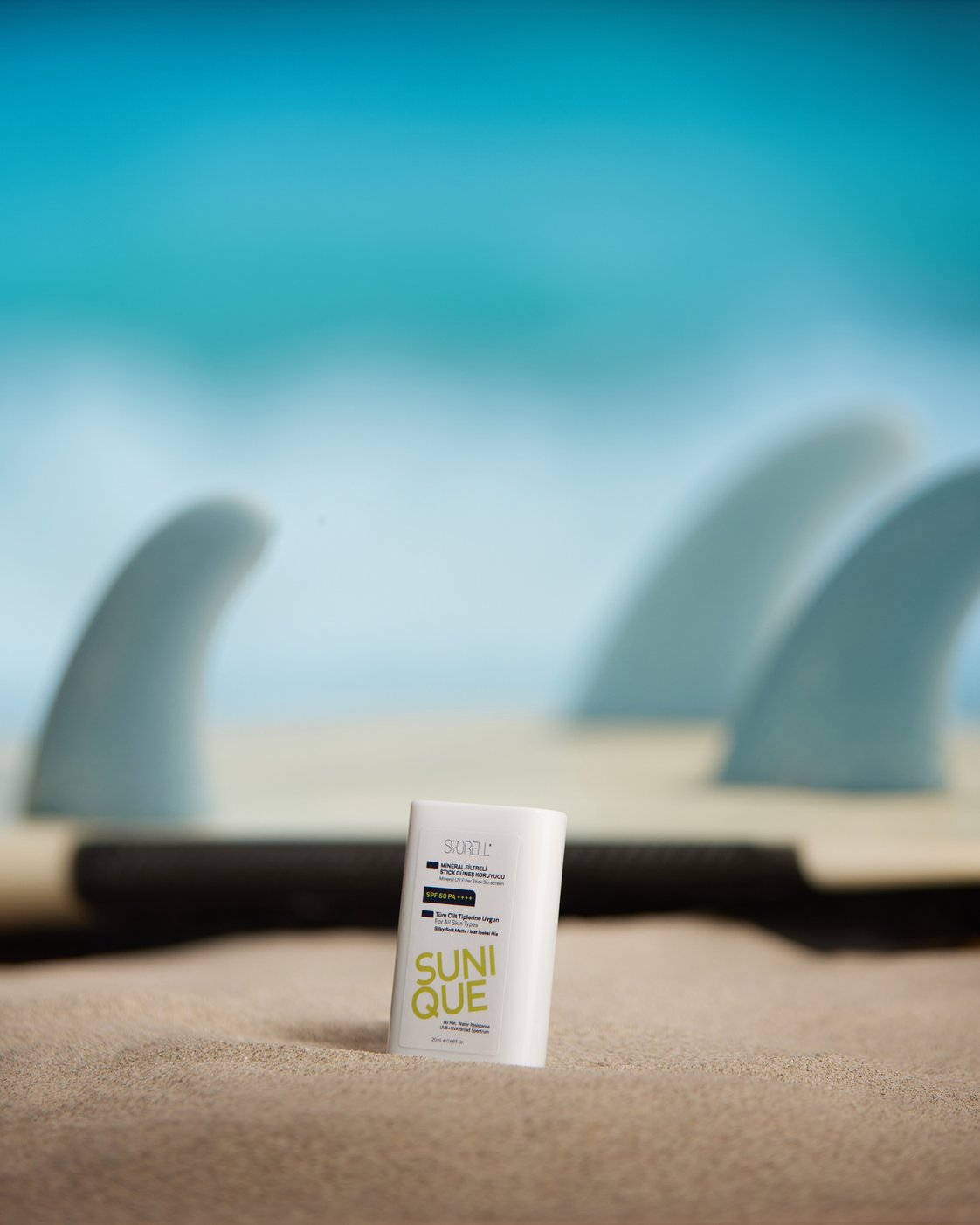 Sunique Stick SPF 50 sunscreen on sand with surfboard fins in the background
