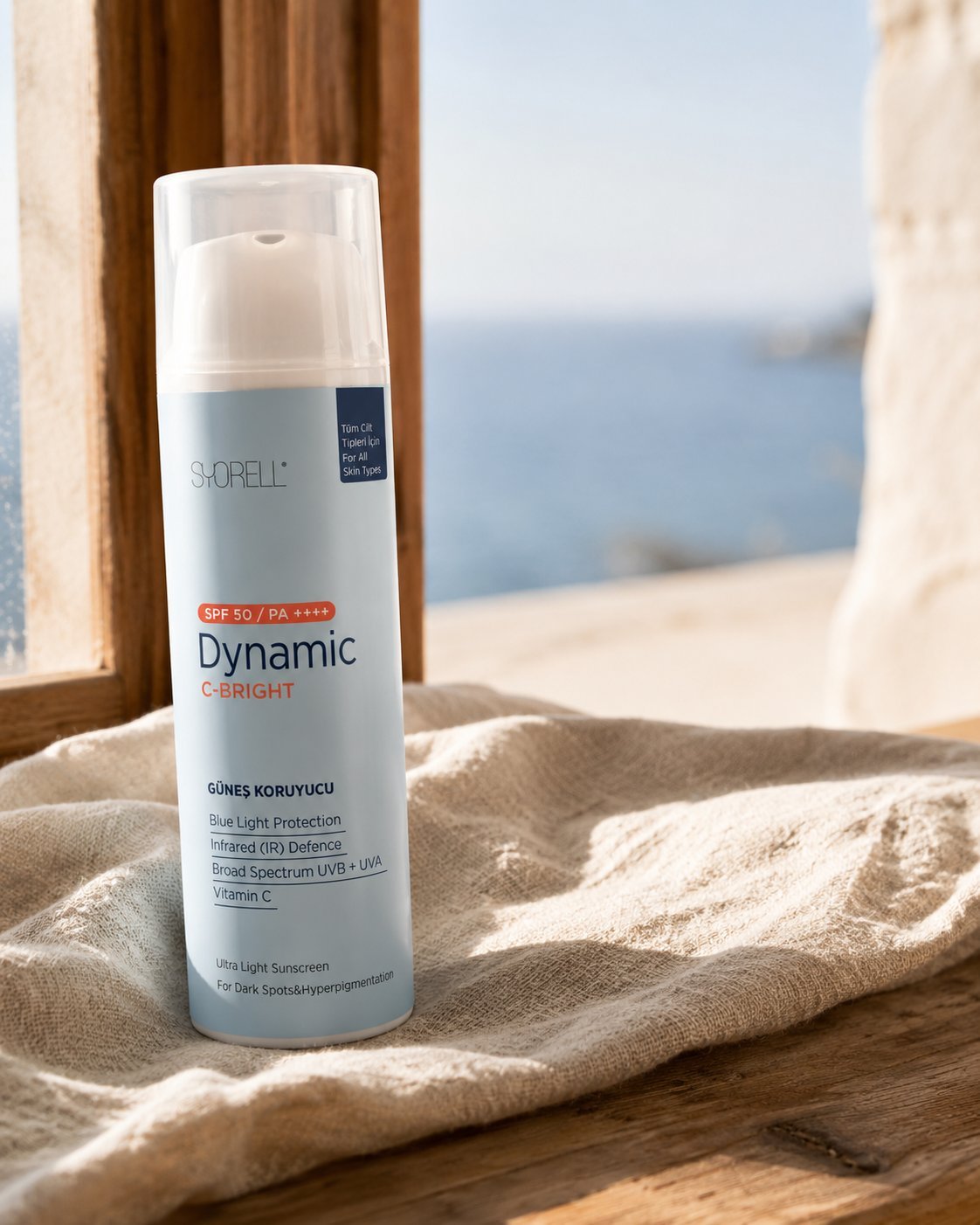 Dynamic C-Bright SPF 50 sunscreen on linen and worn wood near an open window with blurred Mediterranean sea view