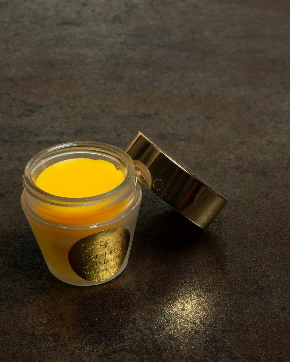 Syorell Restorative Balm in frosted glass jar with gold lid, amber balm visible inside, on dark slate surface